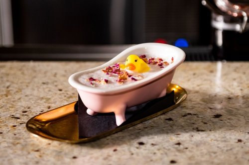A pink bathtub-shaped dish filled with a creamy dessert, topped with rose petals and a rubber duck.