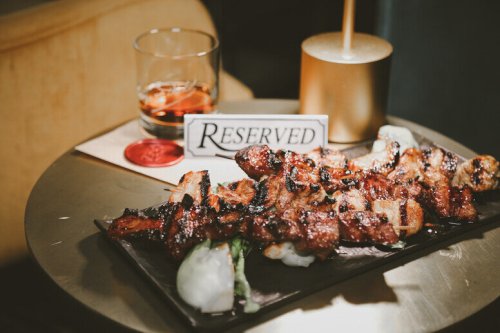 A plate of grilled skewers on a table with a reserved sign and a glass of whiskey.