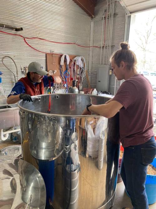 Two individuals work together over a large stainless steel tank in a workshop, focused on their task.