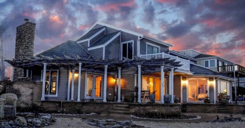 A coastal home with a wraparound porch, stone chimney, and large windows, set against a colorful sunset sky.