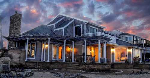 A coastal home with a wraparound porch, stone chimney, and large windows, set against a colorful sunset sky.