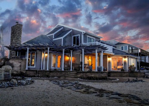 A coastal home at dusk, featuring a stone chimney, porch, and warm lights glowing from the windows against a colorful sky.