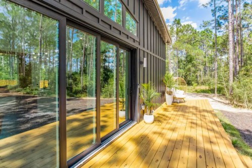 A modern home with large glass windows and a wooden deck, surrounded by trees and greenery.