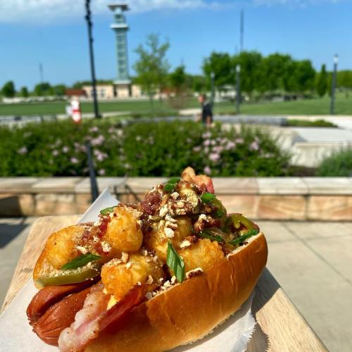 A loaded hot dog topped with tater tots, bacon, and green onions, served on a wooden board with a park in the background.