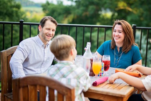 A smiling couple enjoys a meal outdoors with a child, surrounded by drinks and a scenic background.
