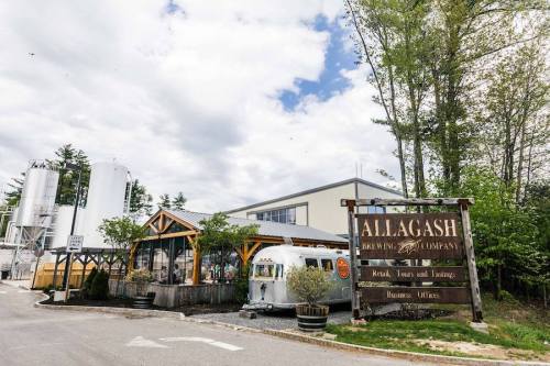 Exterior of Allagash Brewing Company with a sign, outdoor seating, and brewing equipment in a scenic setting.