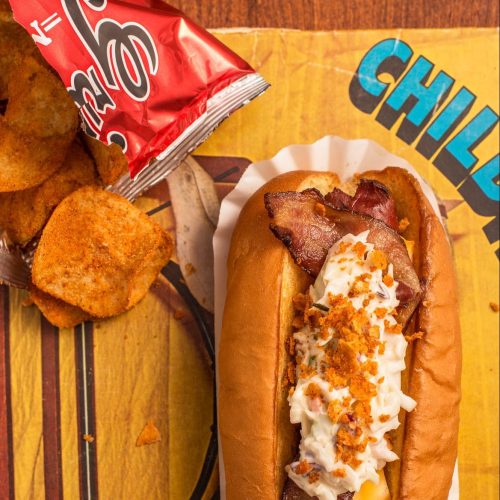 A hot dog topped with bacon and mayonnaise, served with a side of potato chips on a colorful background.