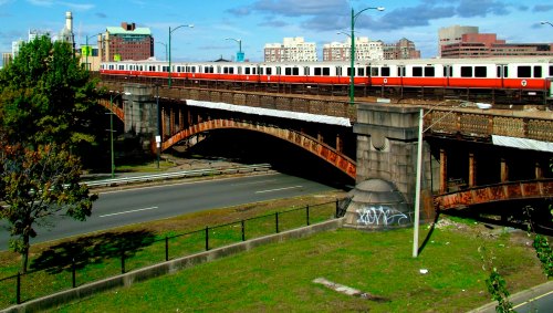 A train crosses a bridge over a highway, with buildings and a blue sky in the background. Green grass is in the foreground.