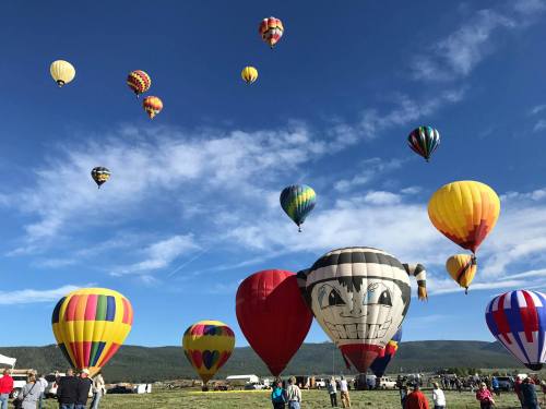 A colorful array of hot air balloons in the sky above a crowd at a festival, with mountains in the background.