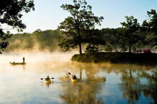Two kayakers paddle on a misty lake at sunrise, with a fisherman in the background and trees lining the shore.