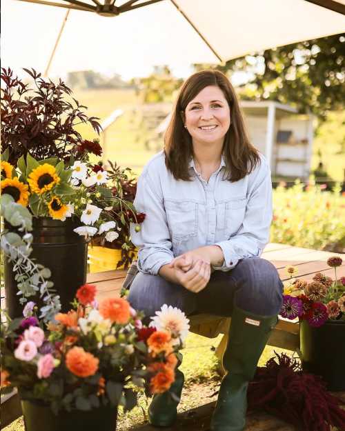 A woman in a light blue shirt and green boots sits among colorful flowers in a garden, smiling under a sunshade.