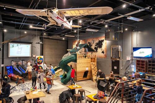 A lively indoor play space with a slide, climbing structure, and a model airplane hanging from the ceiling. Families are enjoying the area.