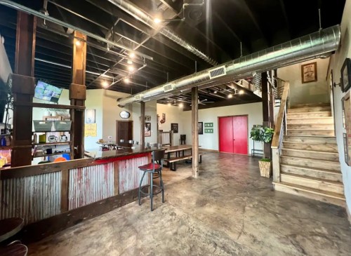Interior of a rustic space featuring a bar, wooden tables, and red doors, with exposed beams and industrial lighting.