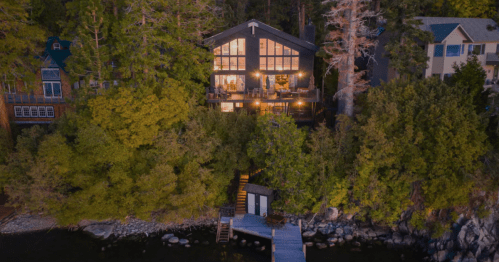 A modern cabin nestled among trees by a lake, featuring large windows and a dock leading to the water.
