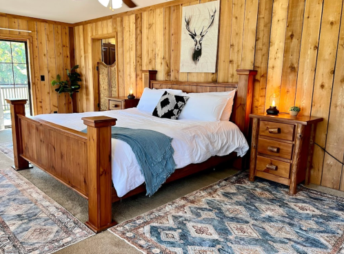 Cozy bedroom with a wooden bed, nightstands, and rustic decor, featuring a deer painting and a patterned rug.