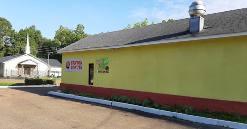 A yellow building with a red base, featuring a sign for "Canyon Donuts," next to a church in the background.