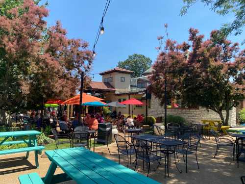 A vibrant outdoor dining area with colorful umbrellas, green trees, and people enjoying their meals on a sunny day.