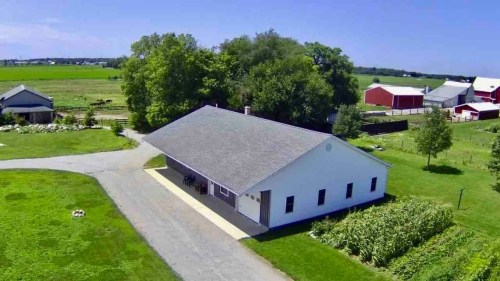 Aerial view of a white farmhouse surrounded by green fields, trees, and red barns under a clear blue sky.