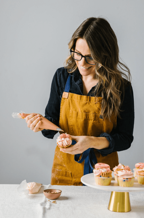 A woman in an apron decorates a cupcake with pink frosting, smiling at her work, with more cupcakes on a table.