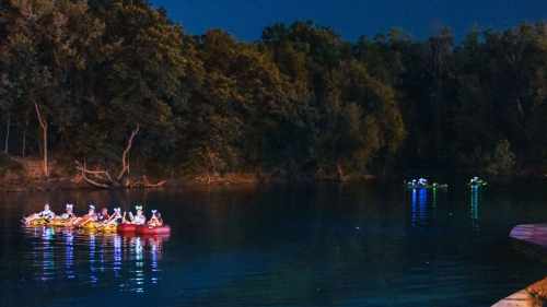 A group of people in kayaks paddles on a calm river at night, surrounded by trees and illuminated by soft lights.
