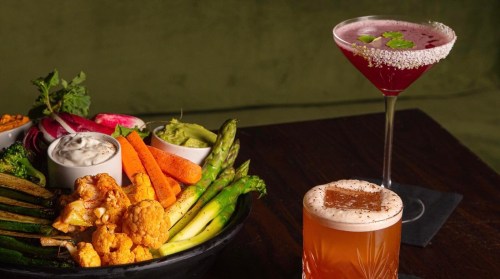 A vibrant platter of fresh vegetables with dips, alongside two colorful cocktails on a wooden table.