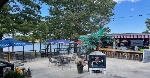 Outdoor bar area by a lake, featuring tables, umbrellas, and a tropical vibe with a sign reading "Best Margaritas."