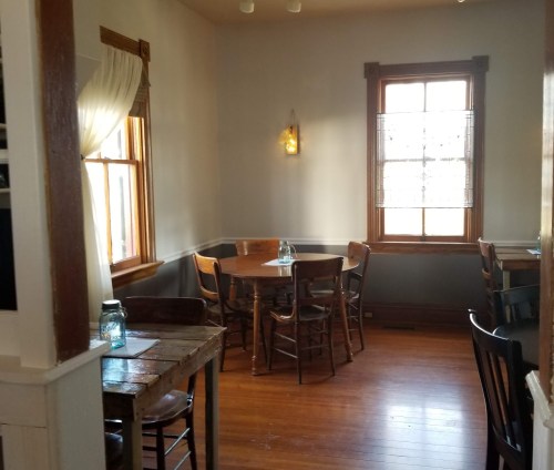 Cozy restaurant interior with wooden tables, chairs, and a large window letting in natural light.