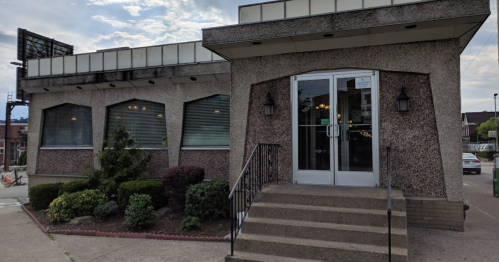 Exterior view of a restaurant with a textured facade, large windows, and a set of stairs leading to the entrance.