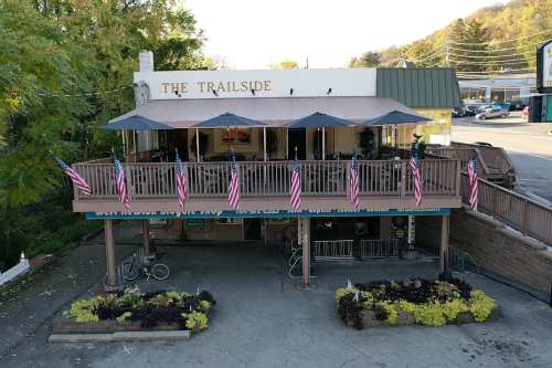 A view of The Trailside building with a deck, umbrellas, and American flags, surrounded by greenery and a parking area.