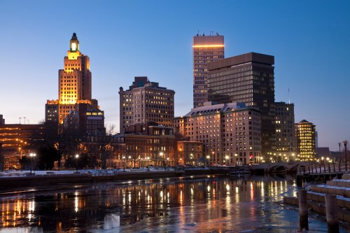 City skyline at dusk, featuring illuminated buildings reflecting in a river with ice along the banks.