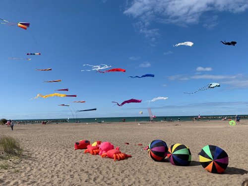 Colorful kites soar in a blue sky over a sandy beach, with vibrant beach balls in the foreground.