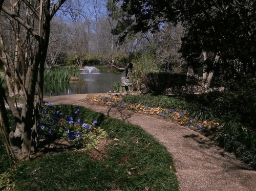 A winding path through a garden with flowers, leading to a pond with a fountain and a statue in the background.