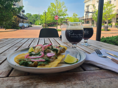 A plate of fish topped with greens and onions, served with lime, alongside two glasses of coffee on a wooden table.