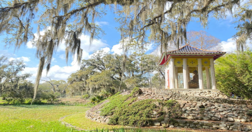 A small, colorful gazebo sits atop a stone mound, surrounded by lush greenery and draped with Spanish moss under a blue sky.