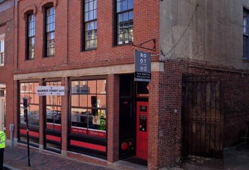 A brick building with large windows, featuring a sign for "Rooted" and "Market Street Eats" on the corner of a street.