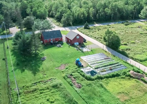 Aerial view of a red farmhouse surrounded by green fields and a garden with rows of crops, near a rural road.