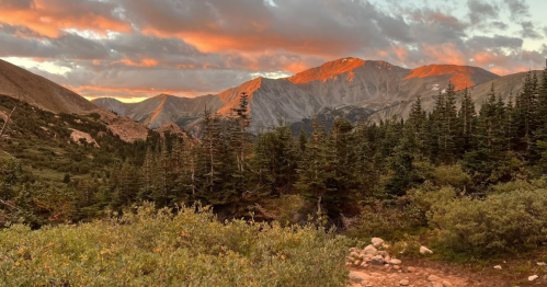 Sunset over mountains with vibrant orange hues, surrounded by lush green trees and rocky terrain.