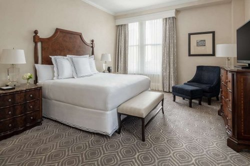 A cozy hotel room featuring a queen bed, a bench, a chair, and elegant decor with natural light from a window.