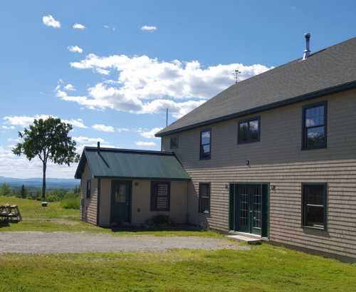 A two-story house with a green roof, surrounded by grass and trees, under a blue sky with scattered clouds.