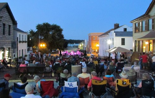 A crowd gathers in a town square for an evening concert, with buildings and trees in the background under a twilight sky.