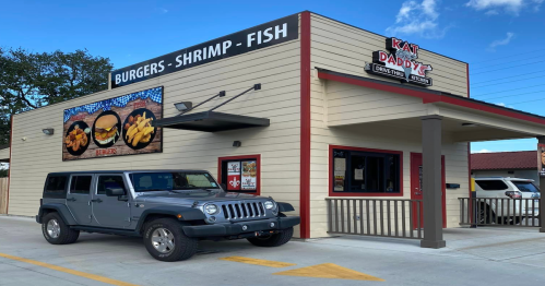 A gray Jeep parked outside a casual restaurant named "Kat Daddy's," featuring a sign for burgers, shrimp, and fish.