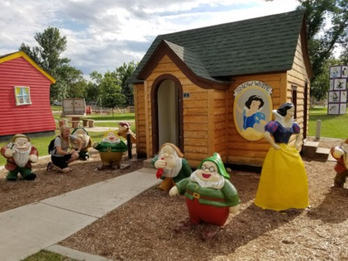 A whimsical scene featuring a small wooden house and colorful dwarf statues, with a figure dressed as Snow White nearby.