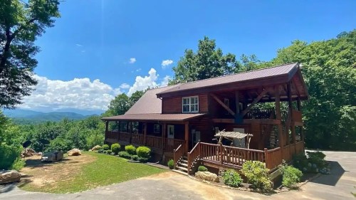 A large wooden cabin surrounded by trees and mountains under a bright blue sky with fluffy clouds.