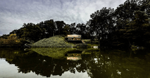 A modern house on a hill, surrounded by trees, reflecting in a calm lake under a cloudy sky.