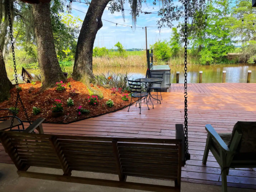 A serene outdoor deck with a swing, flowers, and a view of a calm waterway surrounded by trees.