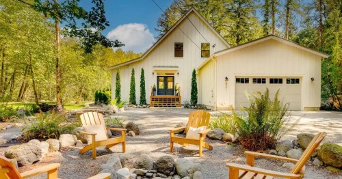 A modern house surrounded by trees, with a gravel driveway and four wooden chairs arranged around a fire pit.