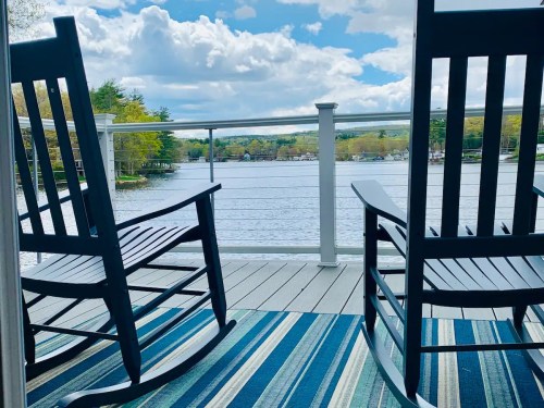 Two rocking chairs on a deck overlooking a calm lake, with a blue sky and fluffy clouds in the background.