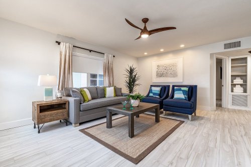 Modern living room with a gray sofa, blue chairs, a coffee table, and decorative plants, featuring bright lighting and neutral tones.