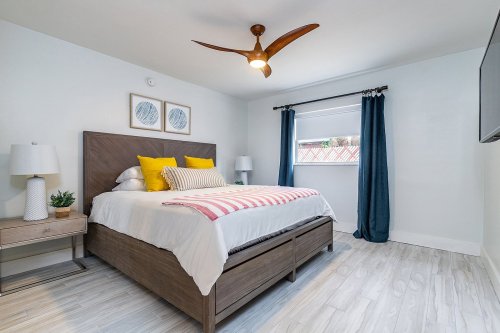 A modern bedroom featuring a bed with yellow and white pillows, a ceiling fan, and blue curtains by a window.