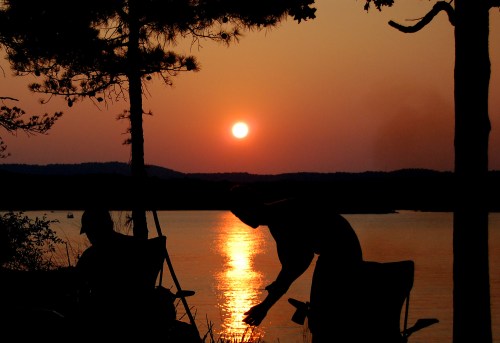 Silhouettes of two people by a lake at sunset, with the sun reflecting on the water and trees in the foreground.